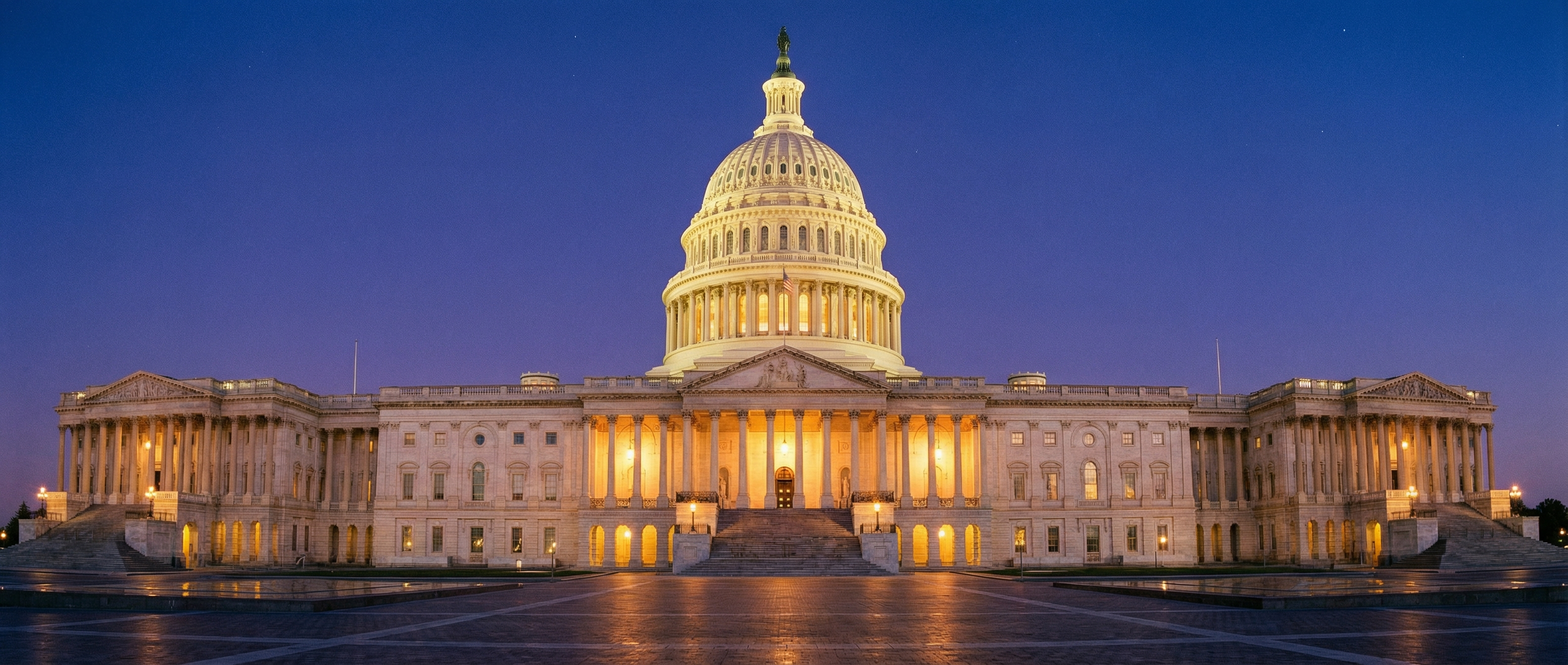 U.S. Capitol Building dome and east front plaza illuminated at twilight by warm amber floodlights against a deep blue-violet sky, marble steps and neoclassical columns gleaming