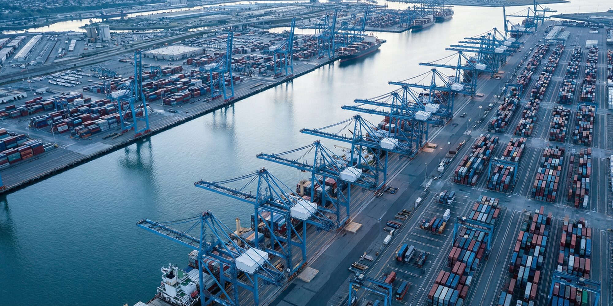 Aerial view of a large U.S. commercial port facility with rows of container cranes stretching along an industrial waterway, steel dock infrastructure in cool blue morning light