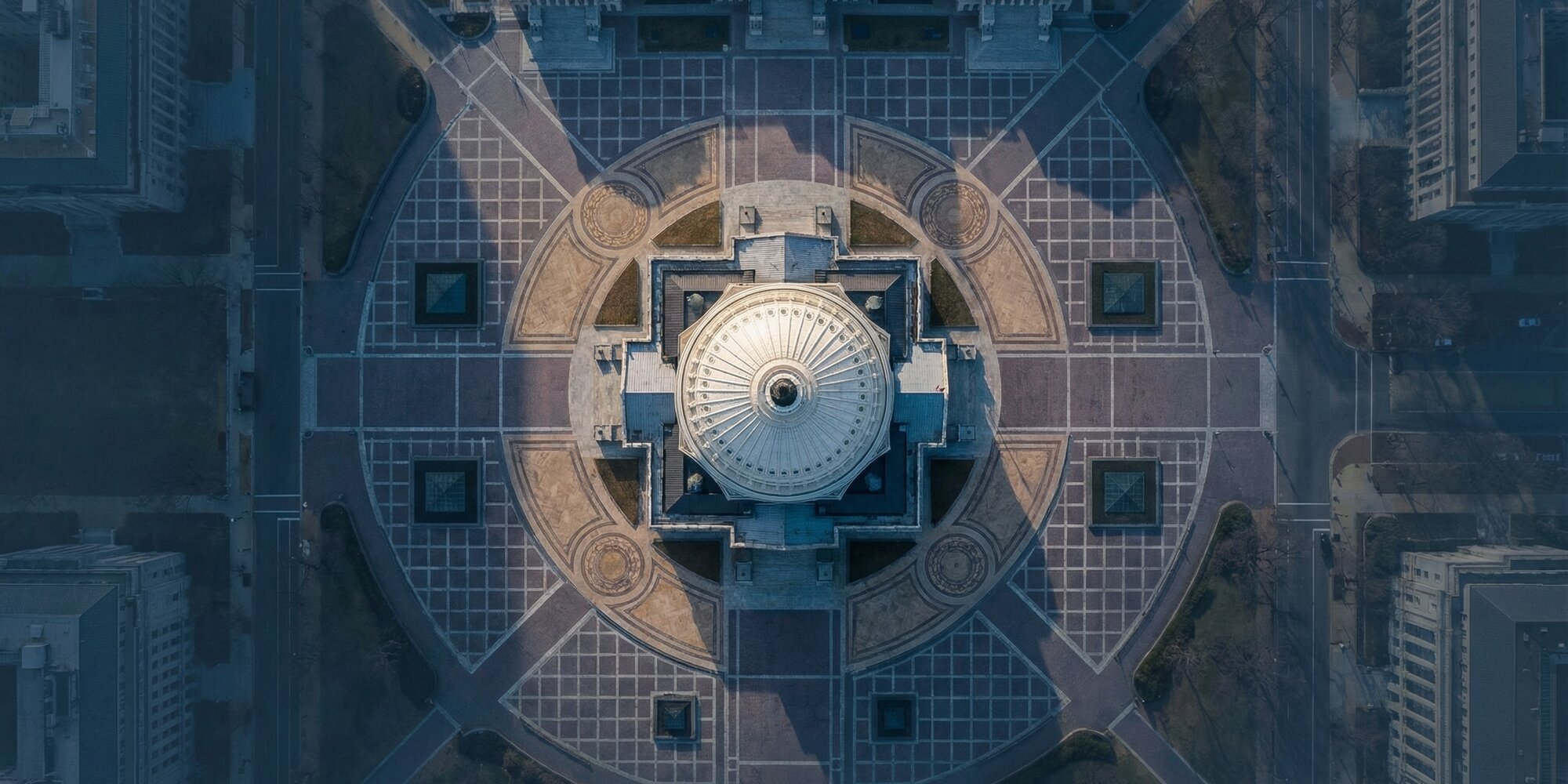 Aerial view looking straight down at the United States Capitol Building rotunda dome surrounded by symmetrical government plaza stonework and geometric pathways in cool dawn light