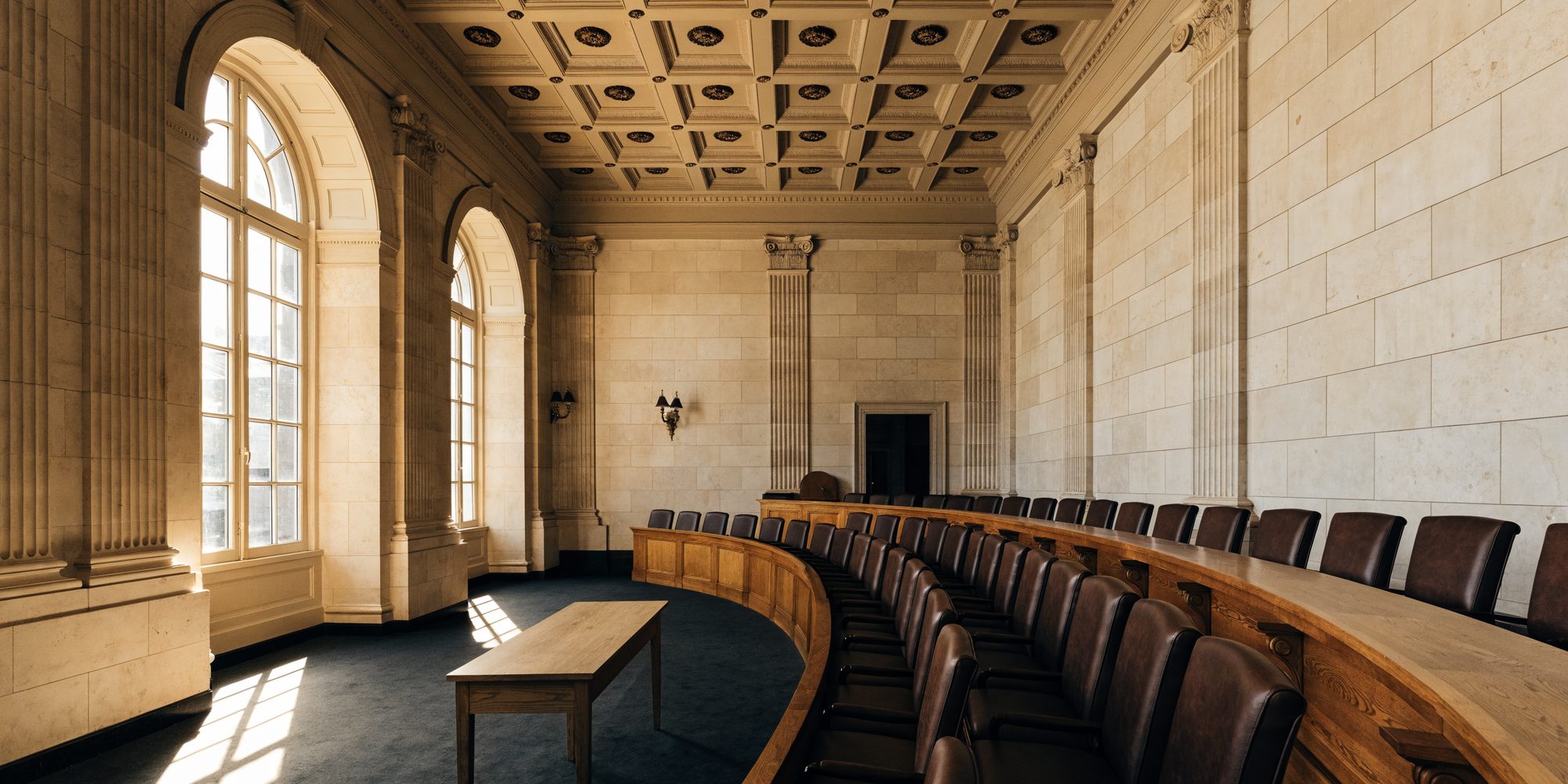 An empty neoclassical committee hearing room, with a long curved dais and rows of leather chairs facing a plain witness table