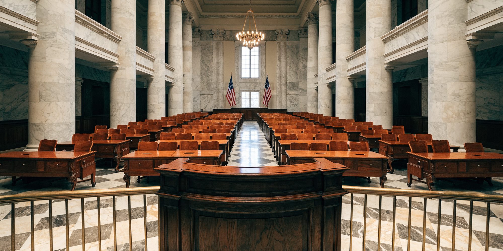 An empty legislative chamber seen from behind the presiding officer's rostrum, looking out over rows of wooden desks