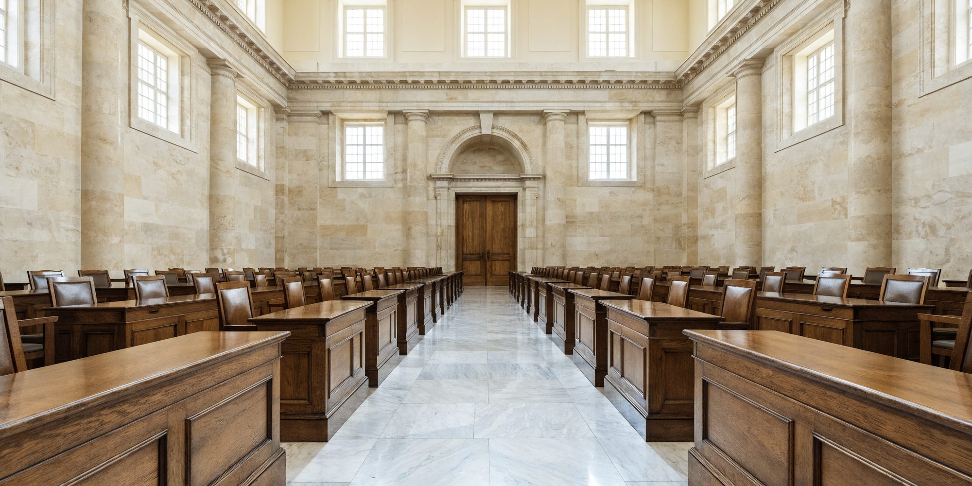 An empty legislative chamber in daylight, looking down the centre aisle toward a set of closed panelled doors