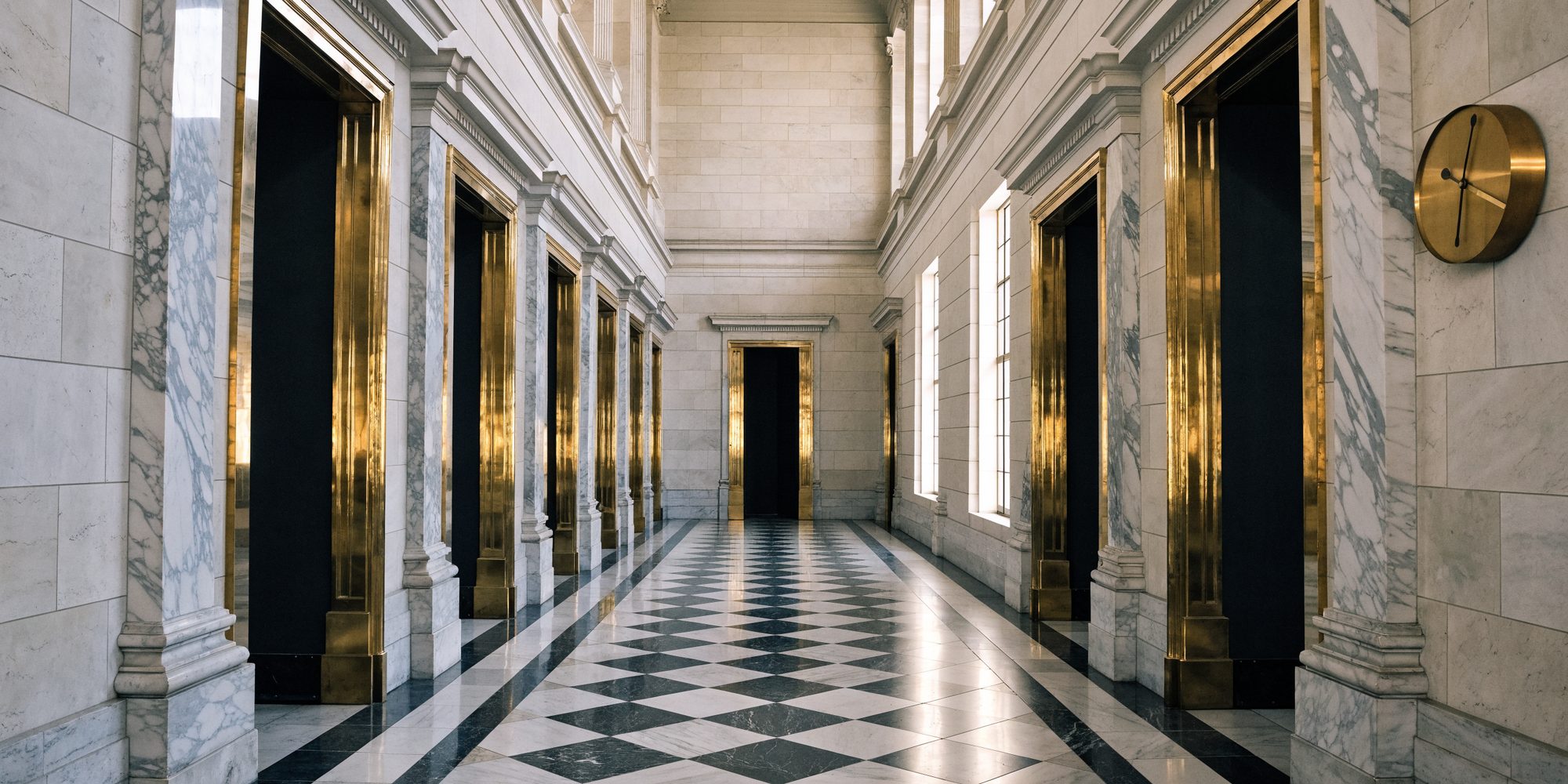 An empty marble capitol corridor lined with brass-trimmed doors, a wall clock on the right