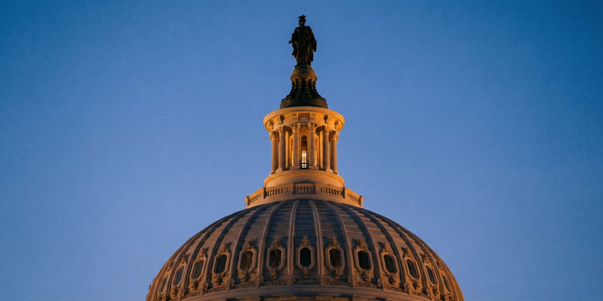The U.S. Capitol dome at dusk, seen from below at a low angle with architectural scroll details lit in amber light against a deep blue evening sky.